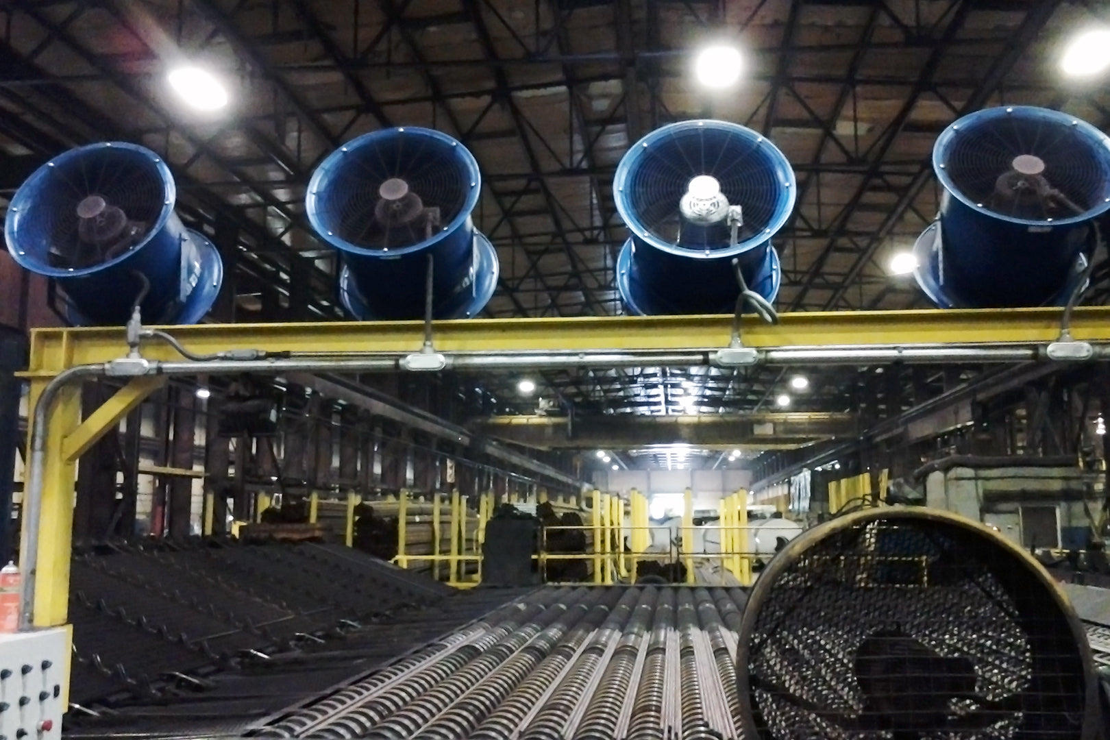 Row of blue 18" industrial high-velocity fans mounted above a production line in a large manufacturing facility to improve airflow and cooling.