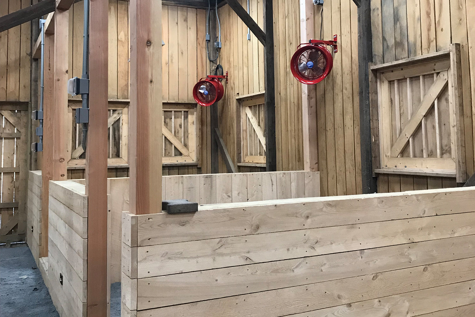 Wooden horse stalls with wall-mounted red circulation fans for barn airflow