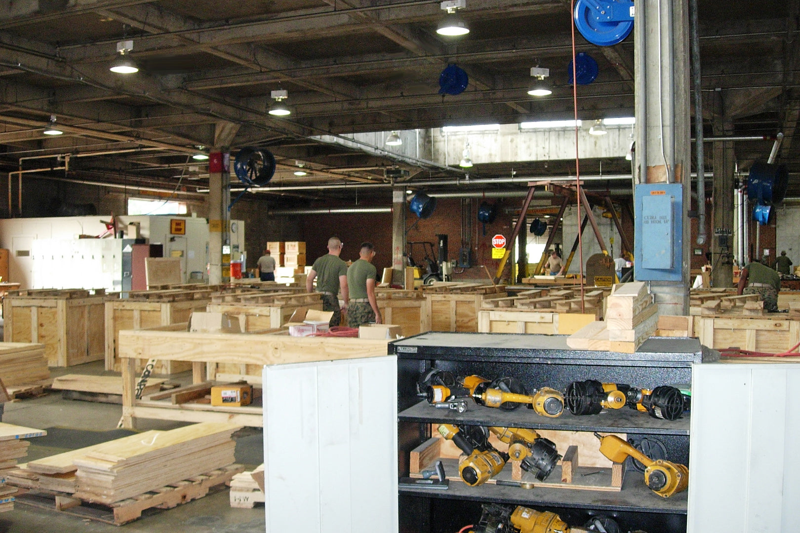 Manufacturing warehouse with military workers assembling wooden crates, power tools, and multiple industrial blue fans mounted overhead