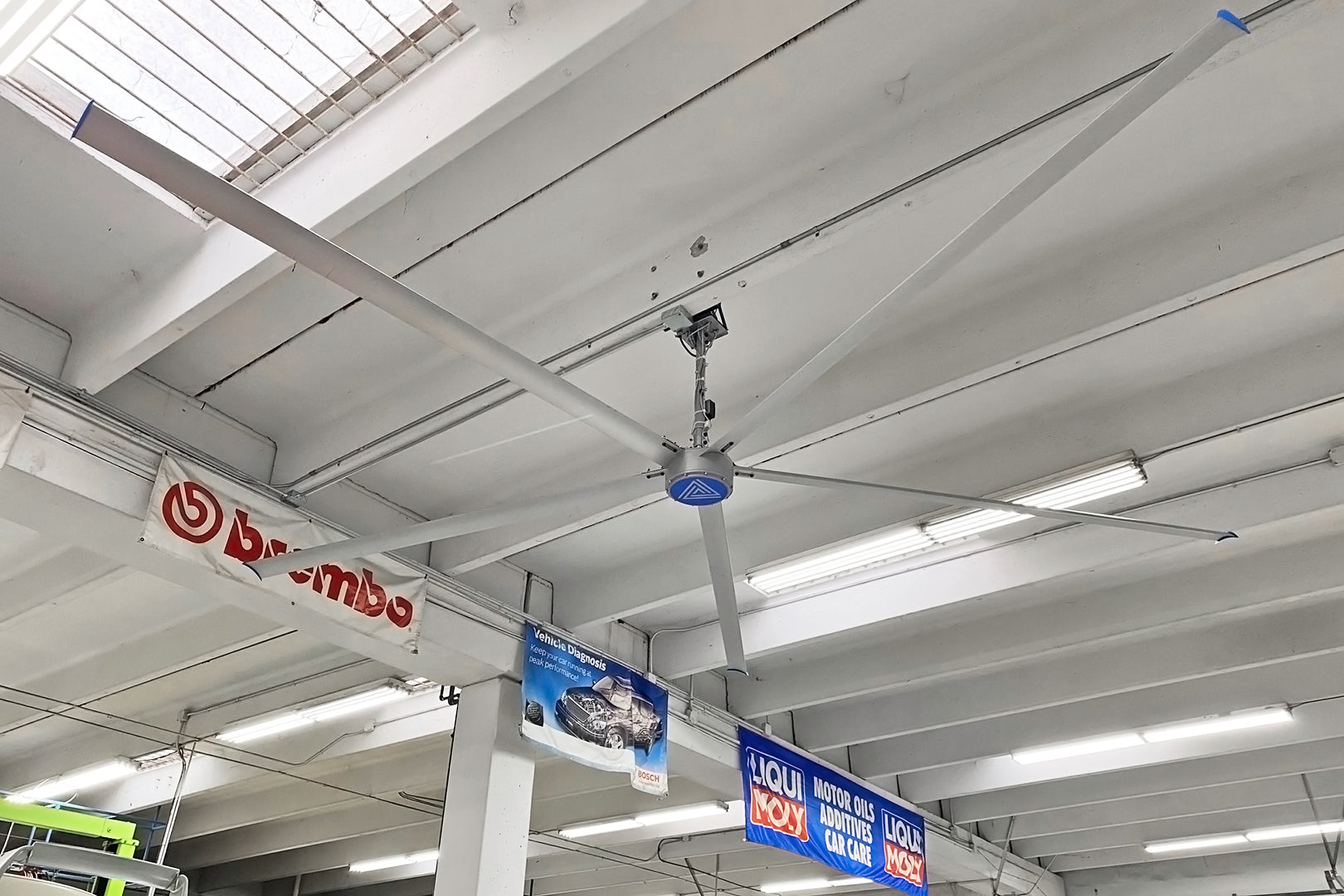 Large industrial HVLS ceiling fan installed in an auto shop with white exposed beams and hanging automotive brand banners