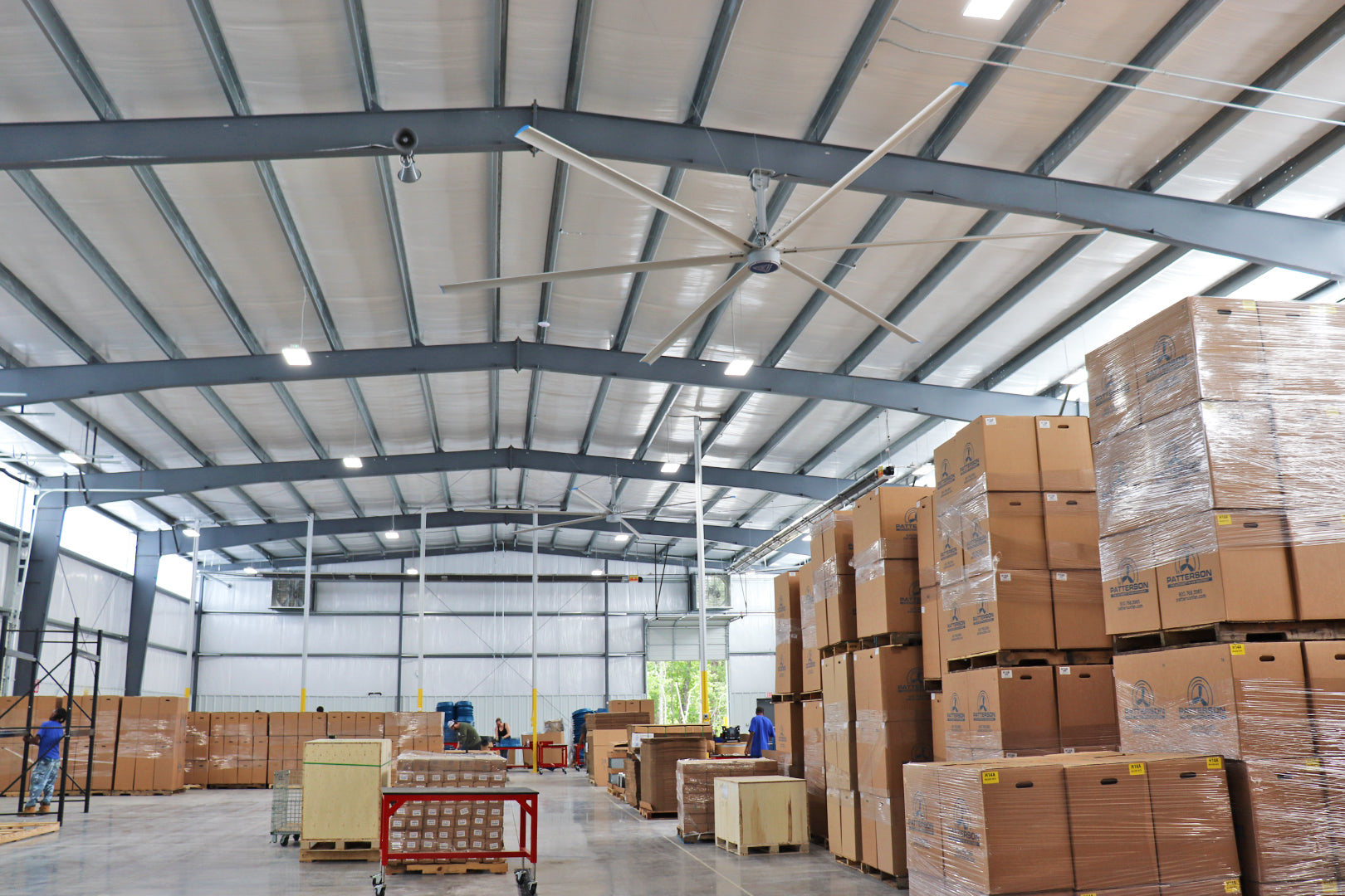 Large industrial ceiling fan operating inside a spacious warehouse with stacked pallets and shipping crates
