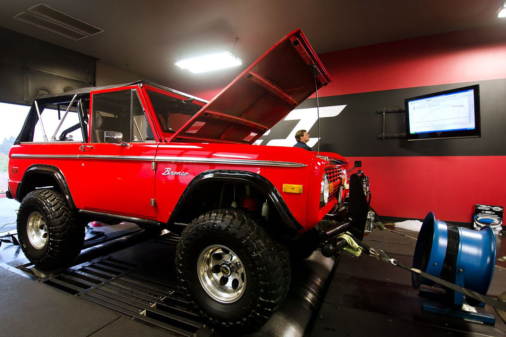 Industrial high-velocity blue barrel fan cooling a red classic Bronco during a dyno test in an automotive tuning shop