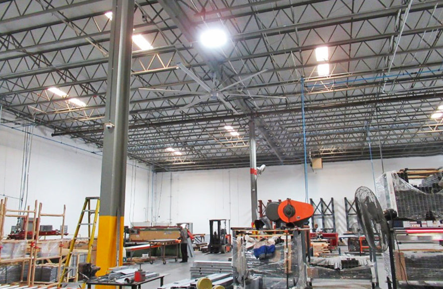 Large industrial ceiling fan operating inside a workspace with metal fabrication equipment, large machinery, and materials organized across the floor.