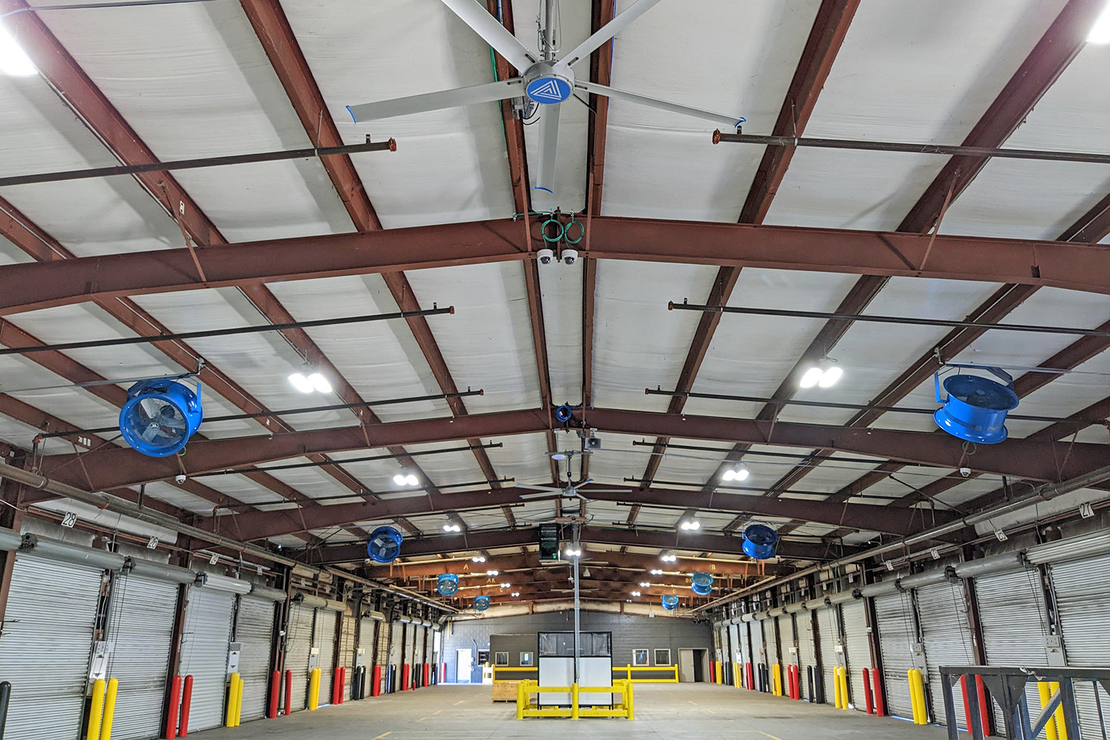 Patterson Fan industrial HVLS ceiling fan and multiple blue barrel fans installed in a warehouse loading dock for improved airflow and ventilation