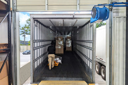 Blue Patterson loading dock fan mounted above an open trailer door providing airflow while a worker loads boxes inside a freight truck