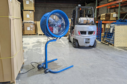 Industrial high-velocity floor fan on a wheeled stand in a warehouse, positioned near a forklift and storage racks