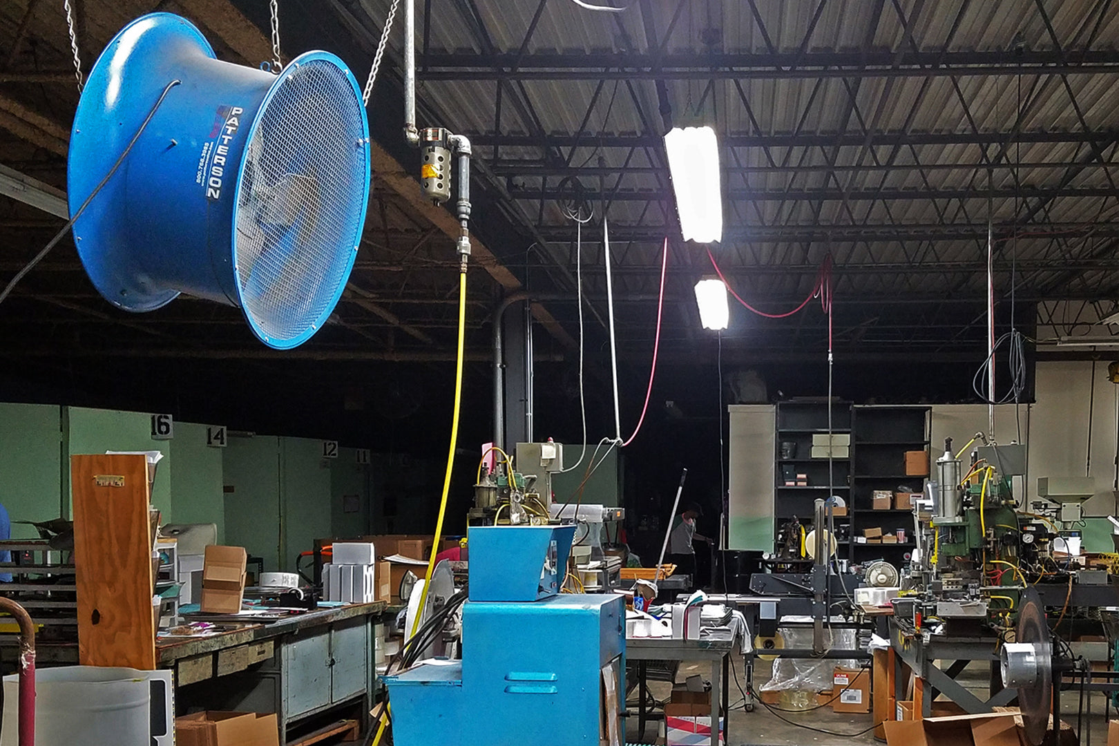 Large industrial hanging ventilation fan suspended from the ceiling above a cluttered workshop with machinery and tools