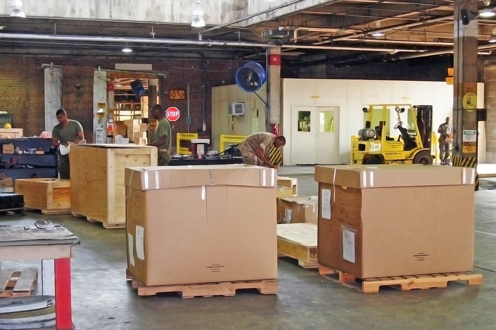 Warehouse shipping area with military workers preparing large boxed pallets and a mounted high-velocity industrial fan in the background