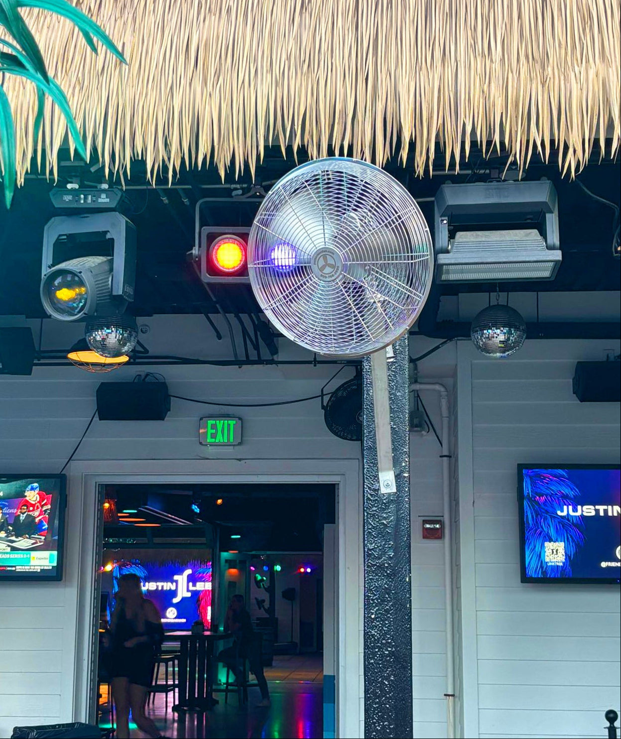A large stainless steel industrial fan mounted on a black support pole outside a bar or club entrance with a thatched roof, stage lights, and screens visible