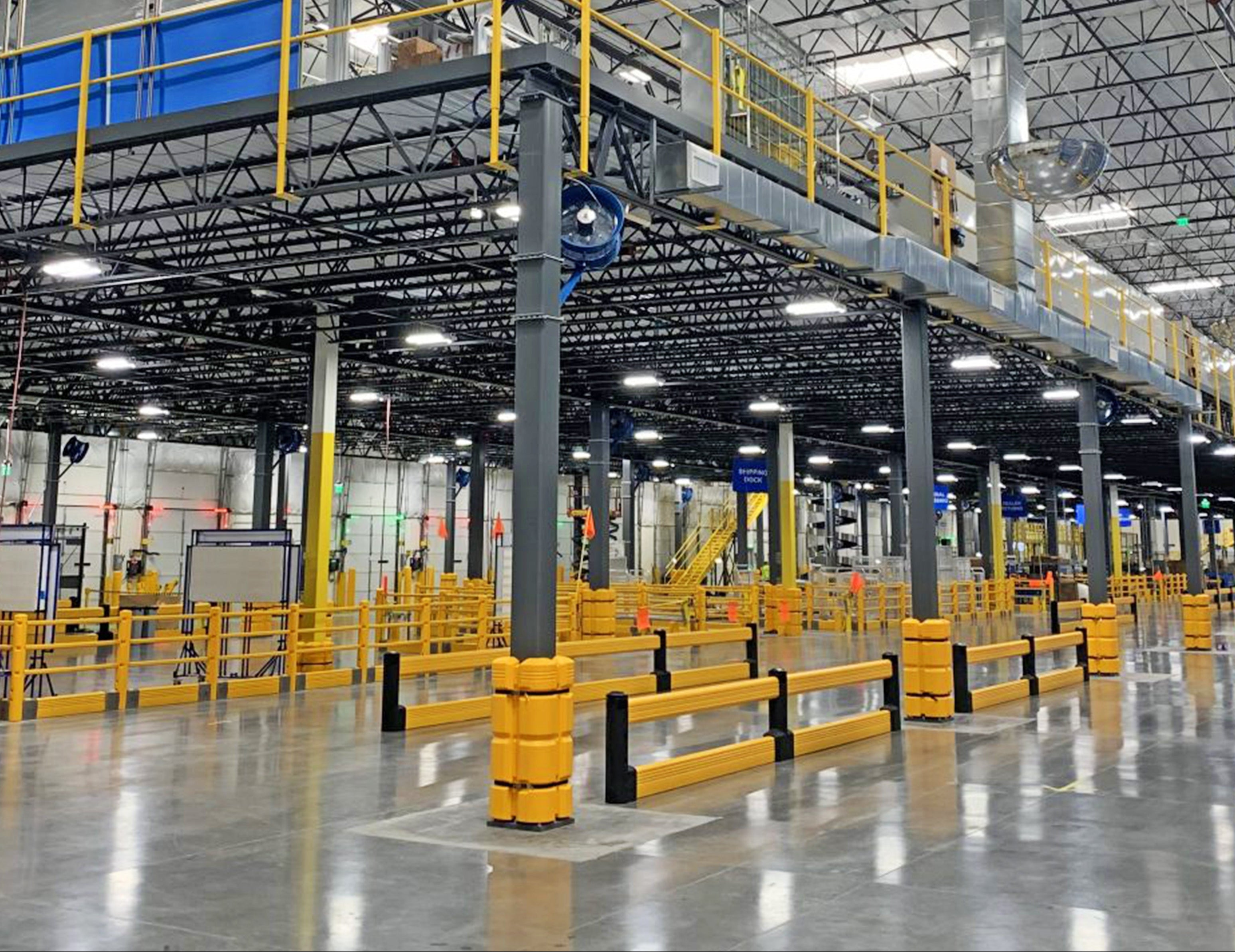 High-velocity industrial wall-mounted fan installed in a large warehouse facility with metal mezzanine, safety railings, and open floor layout