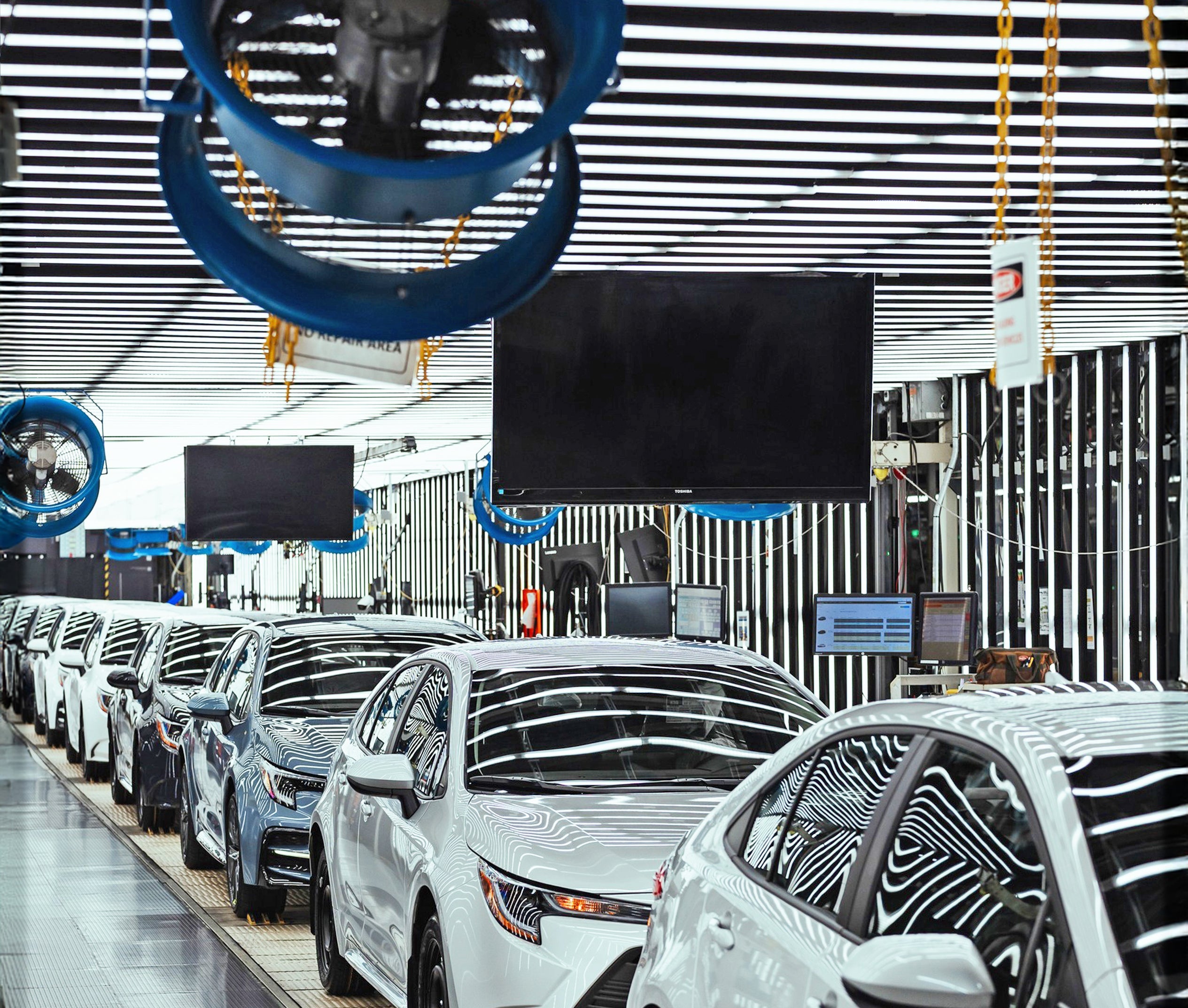 Blue industrial overhead fans providing airflow along an automotive assembly line with new vehicles in production