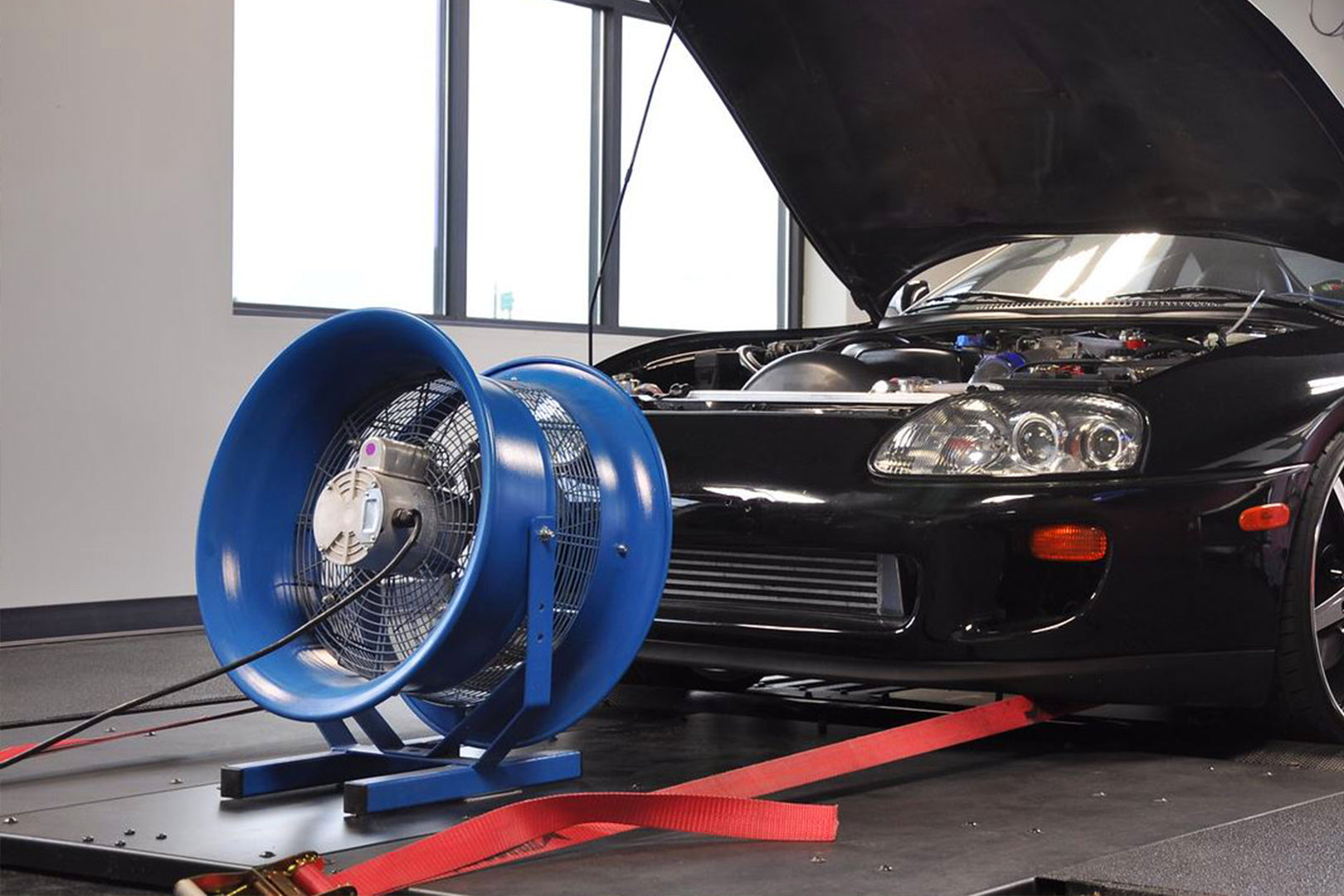 High-power blue industrial shop fan positioned in front of a black sports car on a dyno with the hood open