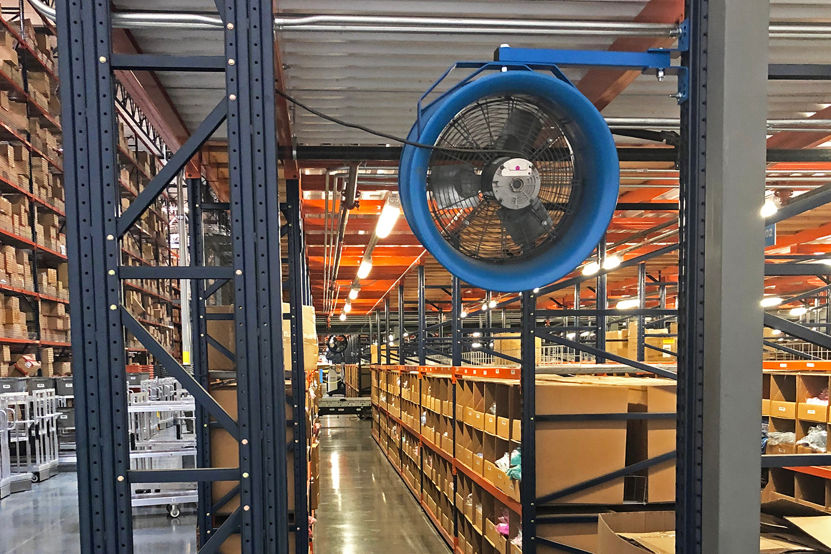 Blue industrial wall-mounted fan in a warehouse aisle with shelving and inventory