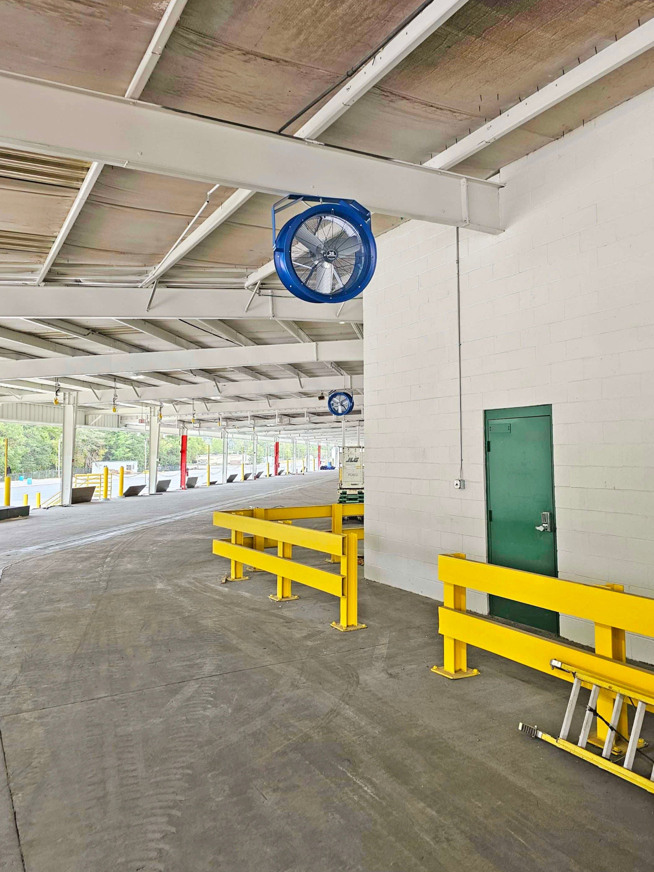 Blue industrial wall-mounted ventilation fan installed in a warehouse loading area with yellow safety rails