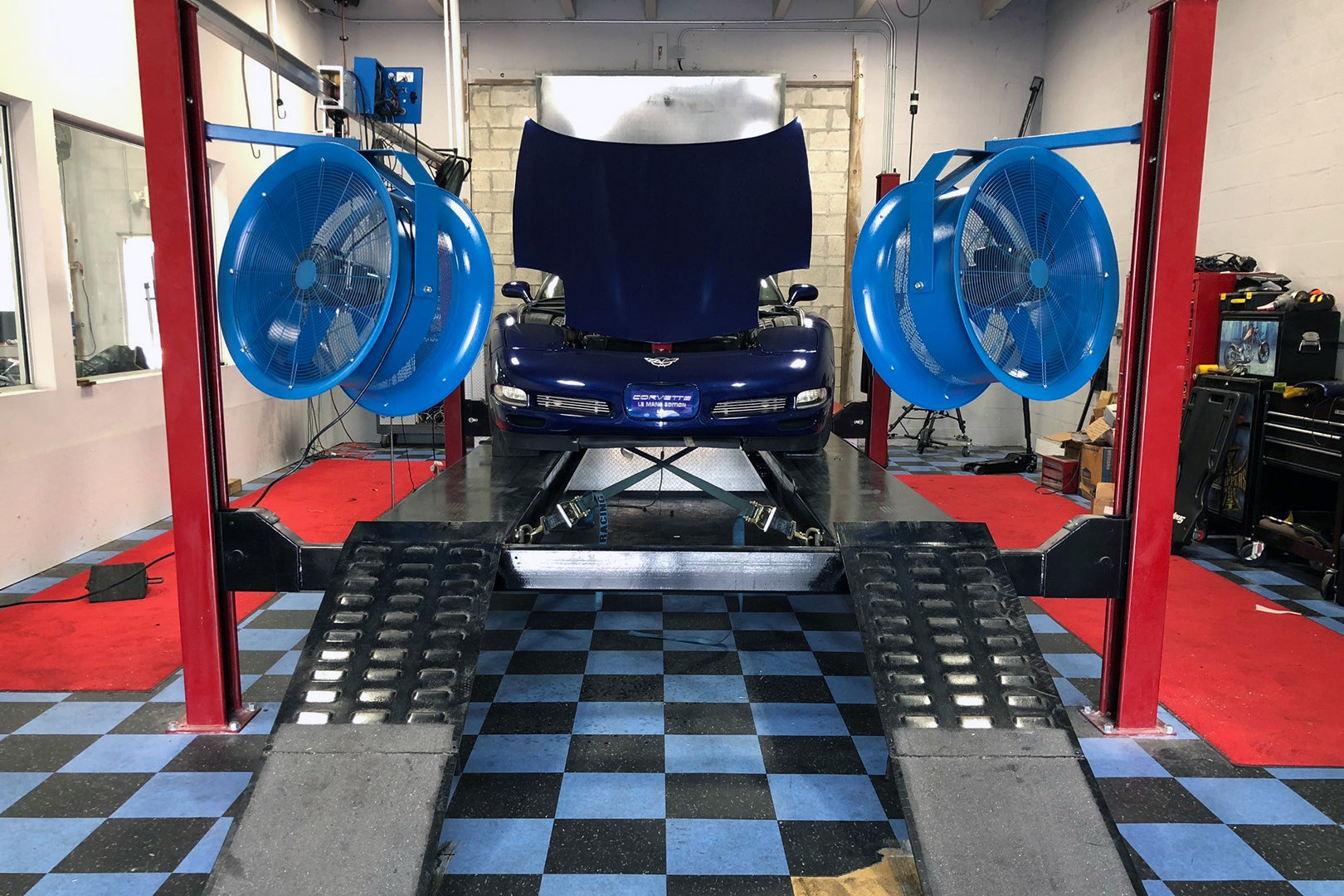 Automotive garage setup with sports care on a life and two large industrial high-velocity blue fans positioned for engine cooling
